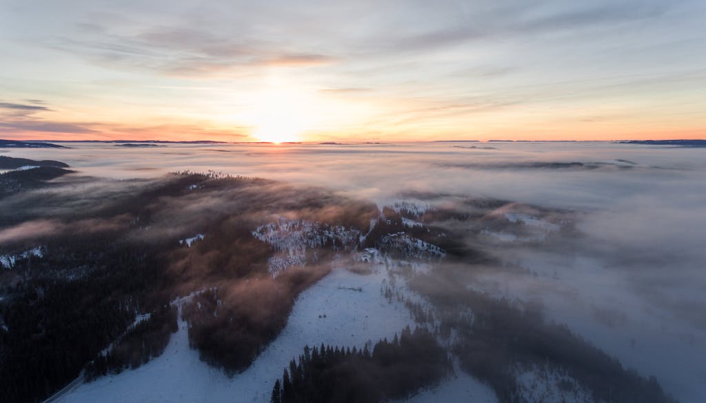 A breathtaking aerial view of a fog-covered winter landscape at sunrise, with snow-dusted hills and dramatic sky.